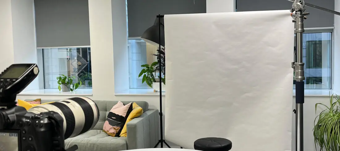 Behind-the-scenes headshot photography setup in a modern office space, showing a Canon camera with a 70-200mm lens in the foreground, a white paper backdrop with a posing stool, an overhead light on a boom arm, a hair light on a stand behind, and a white reflector clamped in position below. A grey sofa with colourful cushions and indoor plants visible in the background.