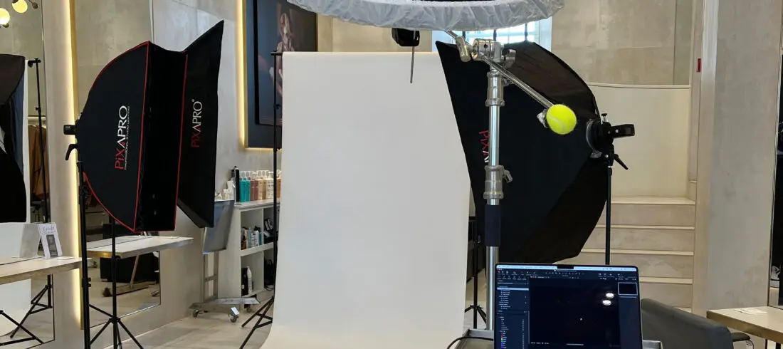 Behind-the-scenes setup of a headshot photography session in an Edinburgh salon, showing a white paper backdrop with two Pixapro softboxes, a large overhead beauty dish on a boom arm with a tennis ball counterweight, a tethered MacBook running Capture One, and a remote trigger on the backdrop.