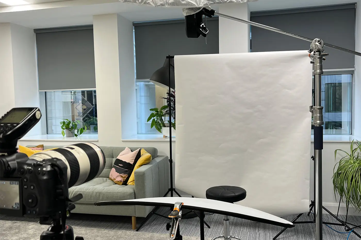 Behind-the-scenes headshot photography setup in a modern office space, showing a Canon camera with a 70-200mm lens in the foreground, a white paper backdrop with a posing stool, an overhead light on a boom arm, a hair light on a stand behind, and a white reflector clamped in position below. A grey sofa with colourful cushions and indoor plants visible in the background.